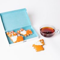 Twist gift box of four hand-iced Princess Corgi gingerbread biscuits beside a cup of tea